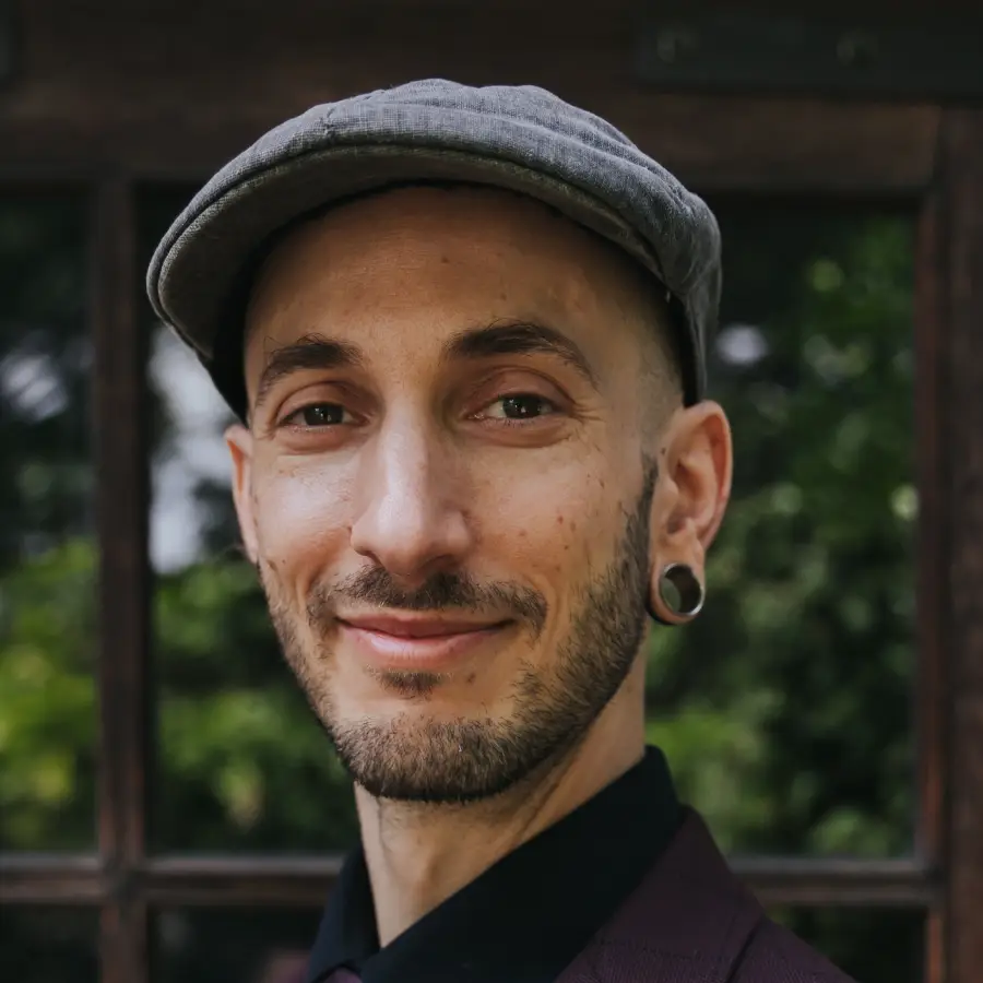 Close-up view of iLan Epstein's face. He has a 5 o'clock shadow on his face, he's making a slight smirk with his mouth, and he's wearing a newsie cap.